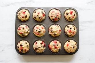 A muffin tin filled with twelve strawberry rhubarb muffins, featuring visible chocolate chips and red fruit pieces, sits on a white marble surface.