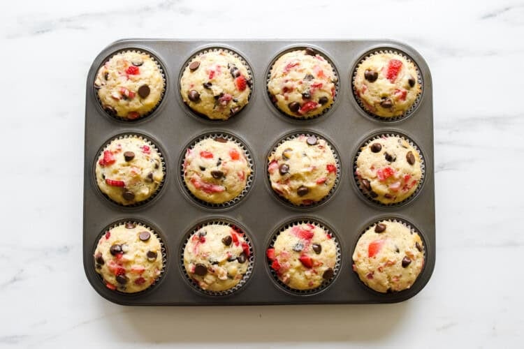 A muffin tin filled with twelve strawberry rhubarb muffins, featuring visible chocolate chips and red fruit pieces, sits on a white marble surface.