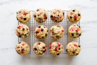 Twelve strawberry rhubarb muffins with chocolate chips and strawberry pieces cooling on a wire rack over a white marble surface.
