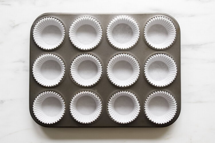 A 12-cup muffin tin with each cup lined with a white paper baking liner, perfect for baking this strawberry rhubarb muffin recipe, placed on a white marble surface.