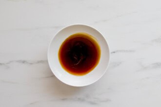 A white bowl filled with brown teriyaki sauce, perfect for Teriyaki Chicken Burgers, sits on a white marble surface.
