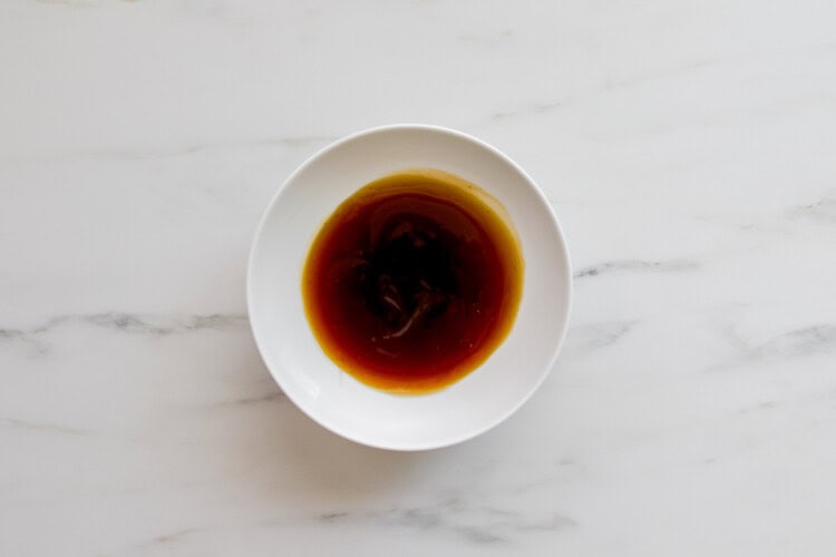A white bowl filled with brown teriyaki sauce, perfect for Teriyaki Chicken Burgers, sits on a white marble surface.
