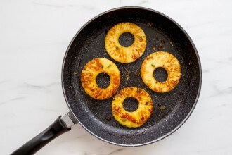 Four pineapple rings being grilled in a black frying pan on a white surface—perfect as a sweet, juicy topping for Teriyaki Chicken Burgers.