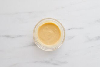 A small glass bowl filled with a creamy, light yellow sauce sits on a white marble surface—perfect for drizzling over Teriyaki Chicken Burgers.