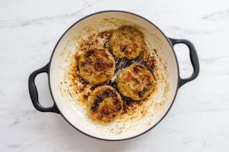 Four Teriyaki Chicken Burgers patties are cooking in a white enameled pan with black handles, showing some char and residue on the pan surface.