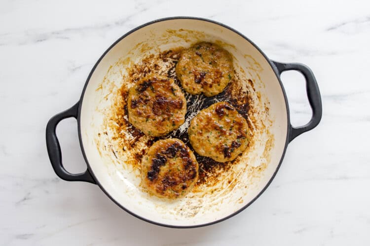 Four Teriyaki Chicken Burgers patties are cooking in a white enameled pan with black handles, showing some char and residue on the pan surface.