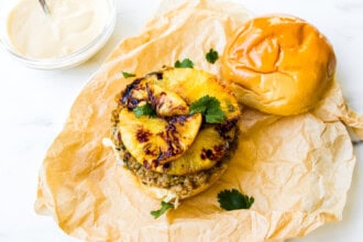 A teriyaki chicken burger patty topped with caramelized pineapple slices and cilantro, sits on a bun with the top half off, displayed on brown parchment paper next to a bowl of sauce.