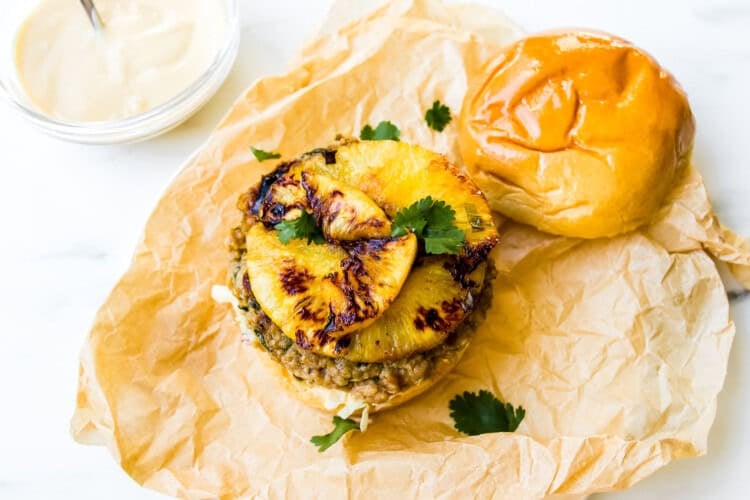 A teriyaki chicken burger patty topped with caramelized pineapple slices and cilantro, sits on a bun with the top half off, displayed on brown parchment paper next to a bowl of sauce.
