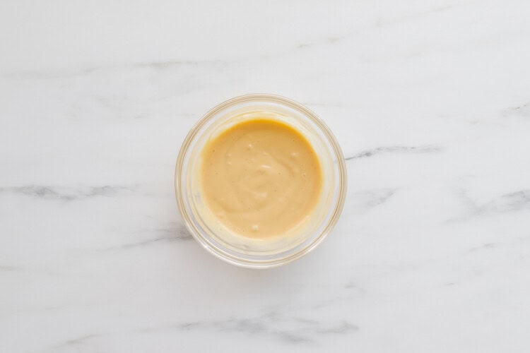 A small glass bowl filled with a creamy, light yellow sauce sits on a white marble surface—perfect for drizzling over Teriyaki Chicken Burgers.