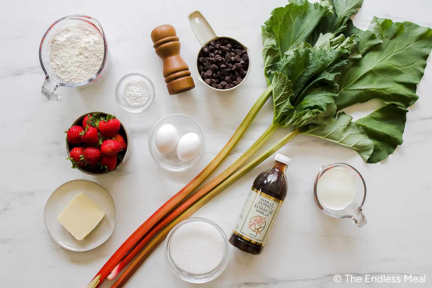 An assortment of baking ingredients, including strawberries, rhubarb, eggs, flour, sugar, butter, milk, vanilla, white chocolate chips—everything you need to create a delicious Strawberry Rhubarb Muffin Cake on your countertop.