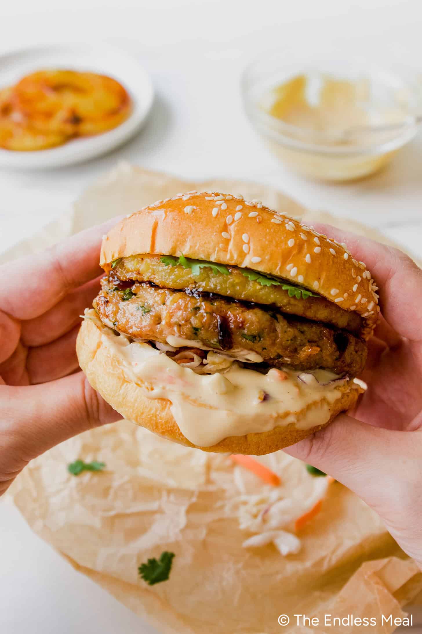 A person holds a sesame seed Teriyaki Chicken Burger with seared patty, caramelized pineapple rings, and teriyaki mayo with a sauce bowl in the background.