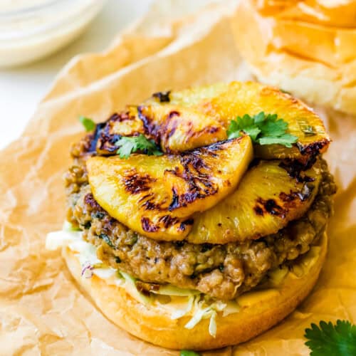 A Teriyaki Chicken Burger with a grilled patty, topped with sweet grilled pineapple slices, shredded cabbage, and fresh cilantro on a bun, served on parchment paper.
