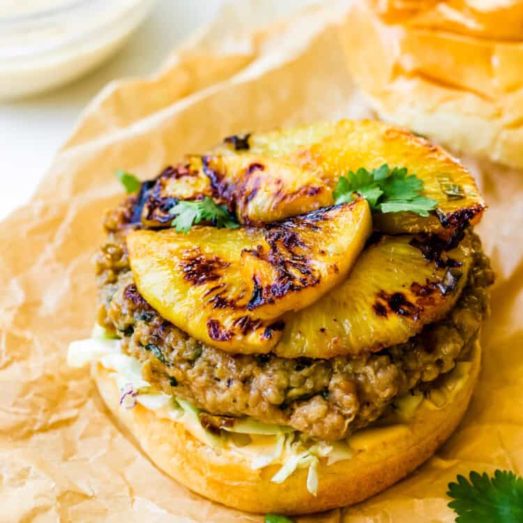 A Teriyaki Chicken Burger with a grilled patty, topped with sweet grilled pineapple slices, shredded cabbage, and fresh cilantro on a bun, served on parchment paper.