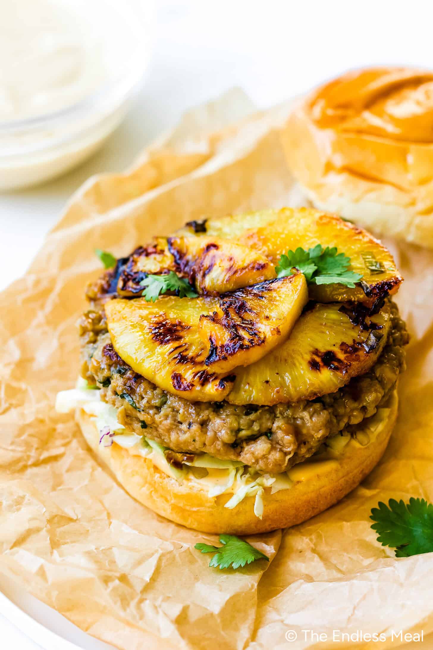 A Teriyaki Chicken Burger topped with caramelized pineapple slices, coleslaw, and cilantro on a toasted bun, set on brown parchment paper.