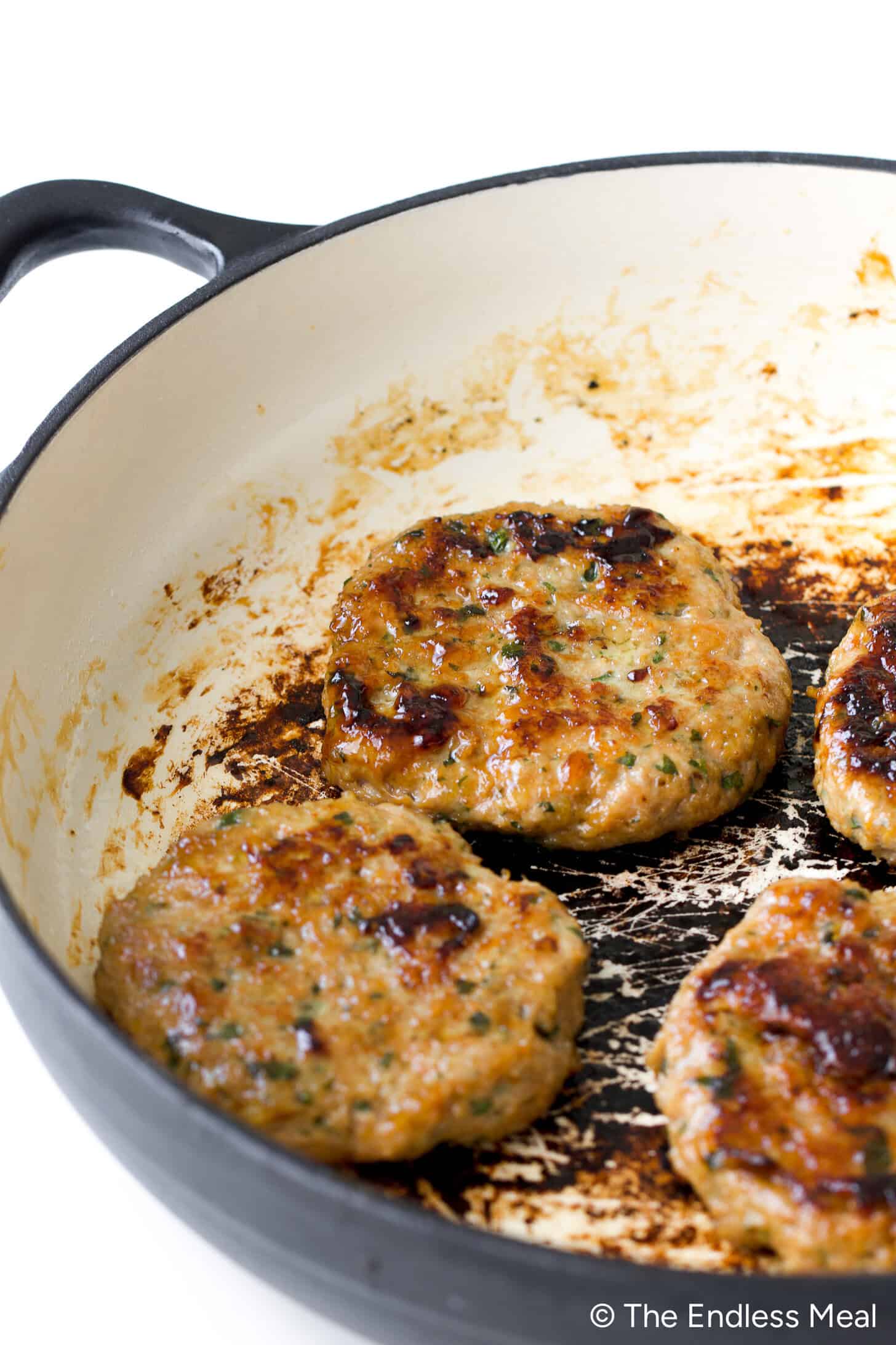Four Teriyaki Chicken Burgers are browning in a white enameled skillet, with visible sear marks and flavorful residue clinging to the pan.