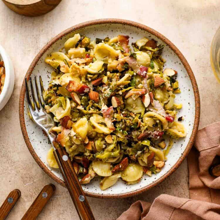 A plate of Roasted Brussels Sprouts Pasta with chopped almonds, greens, and bacon bits is served with a fork and knife on a beige table next to a pink napkin and small bowls of cheese and nuts.
