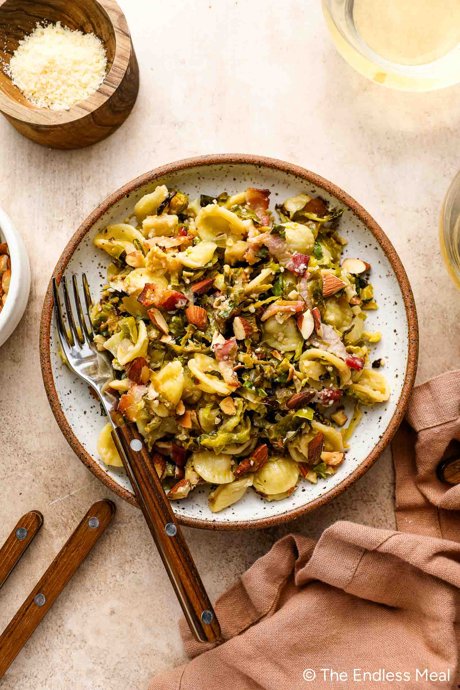 A plate of Roasted Brussels Sprouts Pasta with chopped almonds, greens, and bacon bits is served with a fork and knife on a beige table next to a pink napkin and small bowls of cheese and nuts.