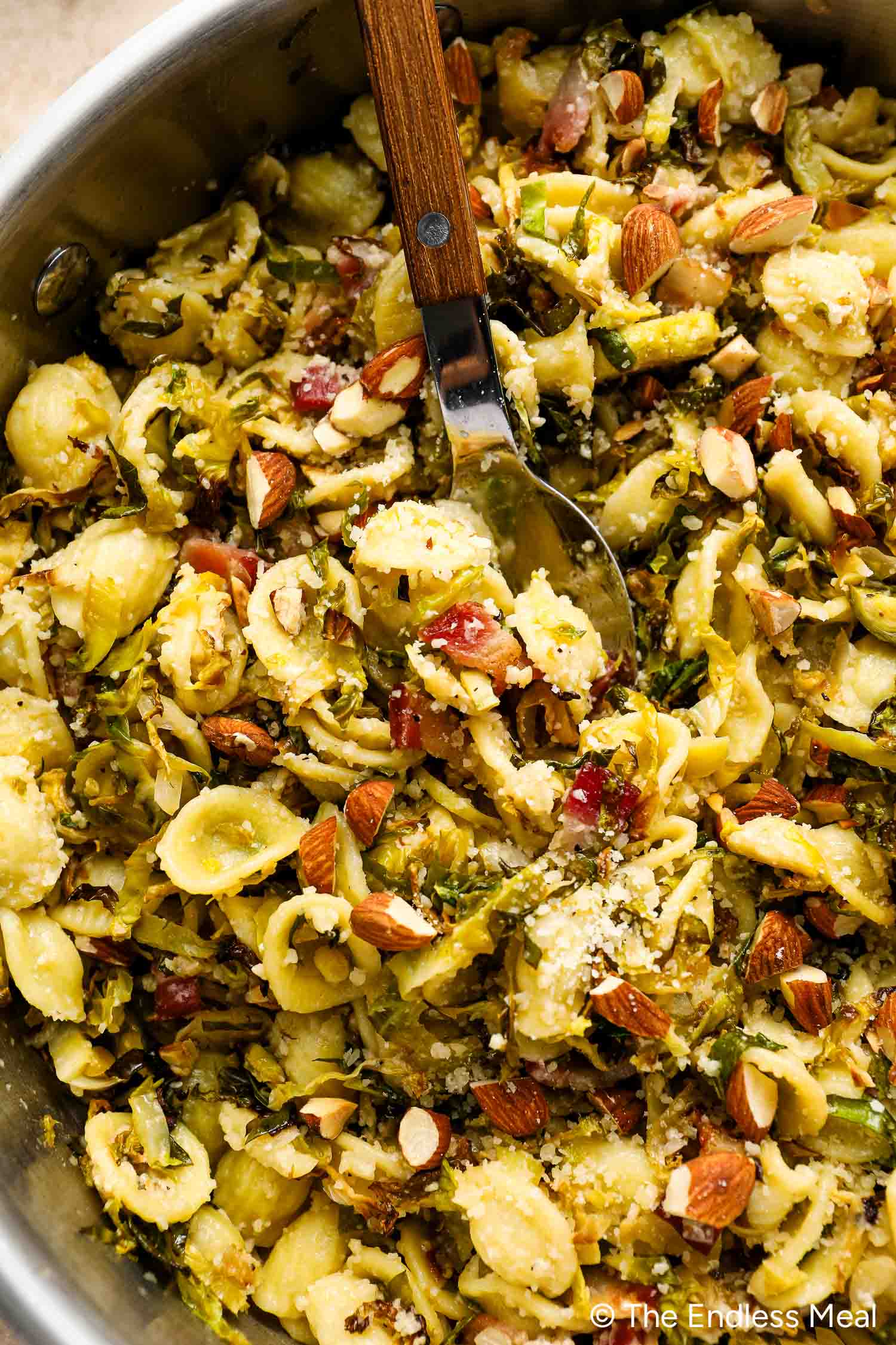A skillet filled with Roasted Brussels Sprouts Pasta—orecchiette, crispy bacon, sliced almonds, grated cheese, and tender roasted Brussels sprouts—all ready to serve with a metal spoon.