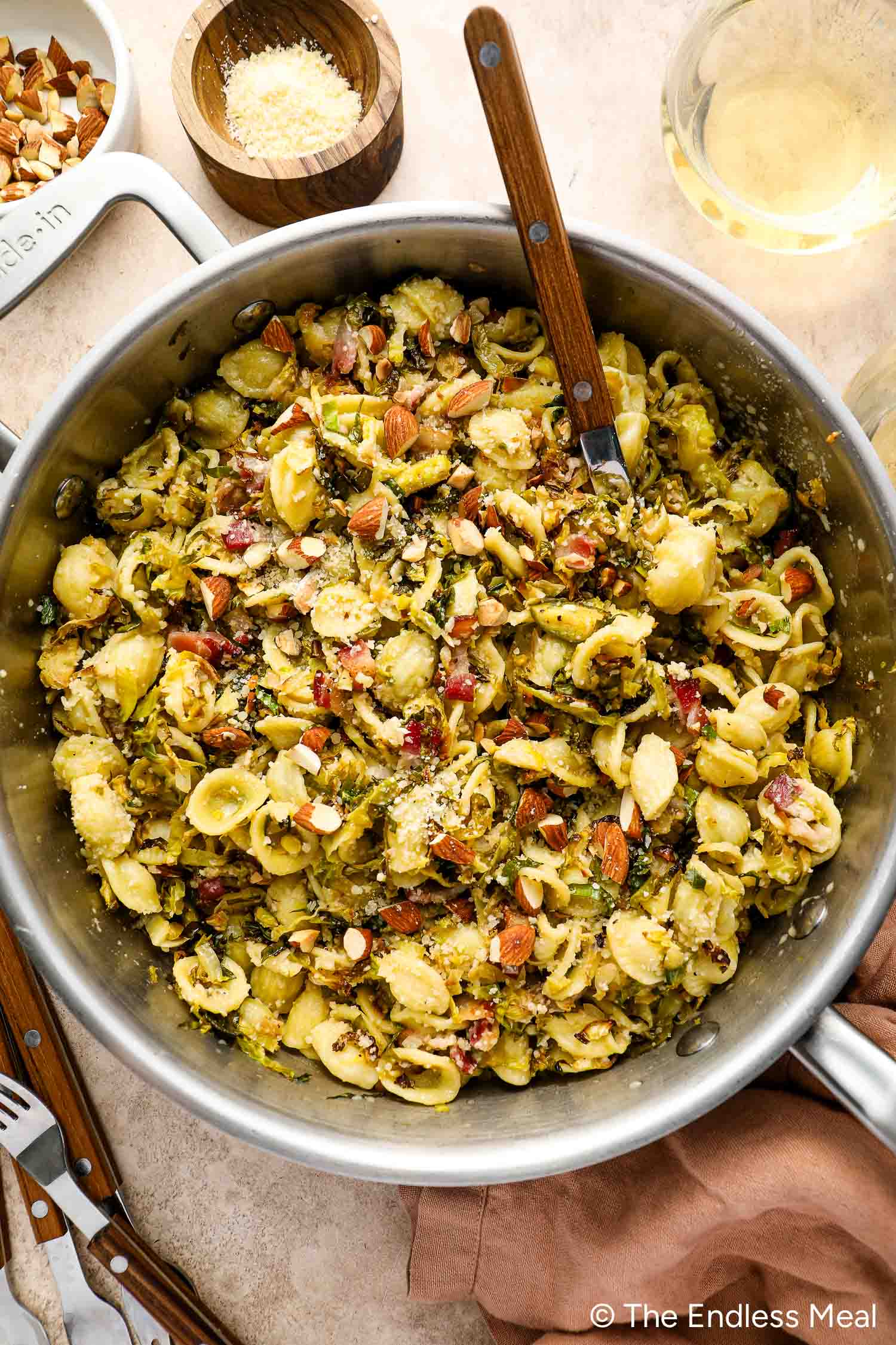 A skillet filled with orecchiette pasta mixed with pesto, chopped almonds, and grated cheese sits next to a bowl of grated cheese and a wooden spoon—perfect for a delicious roasted Brussels sprouts pasta night.
