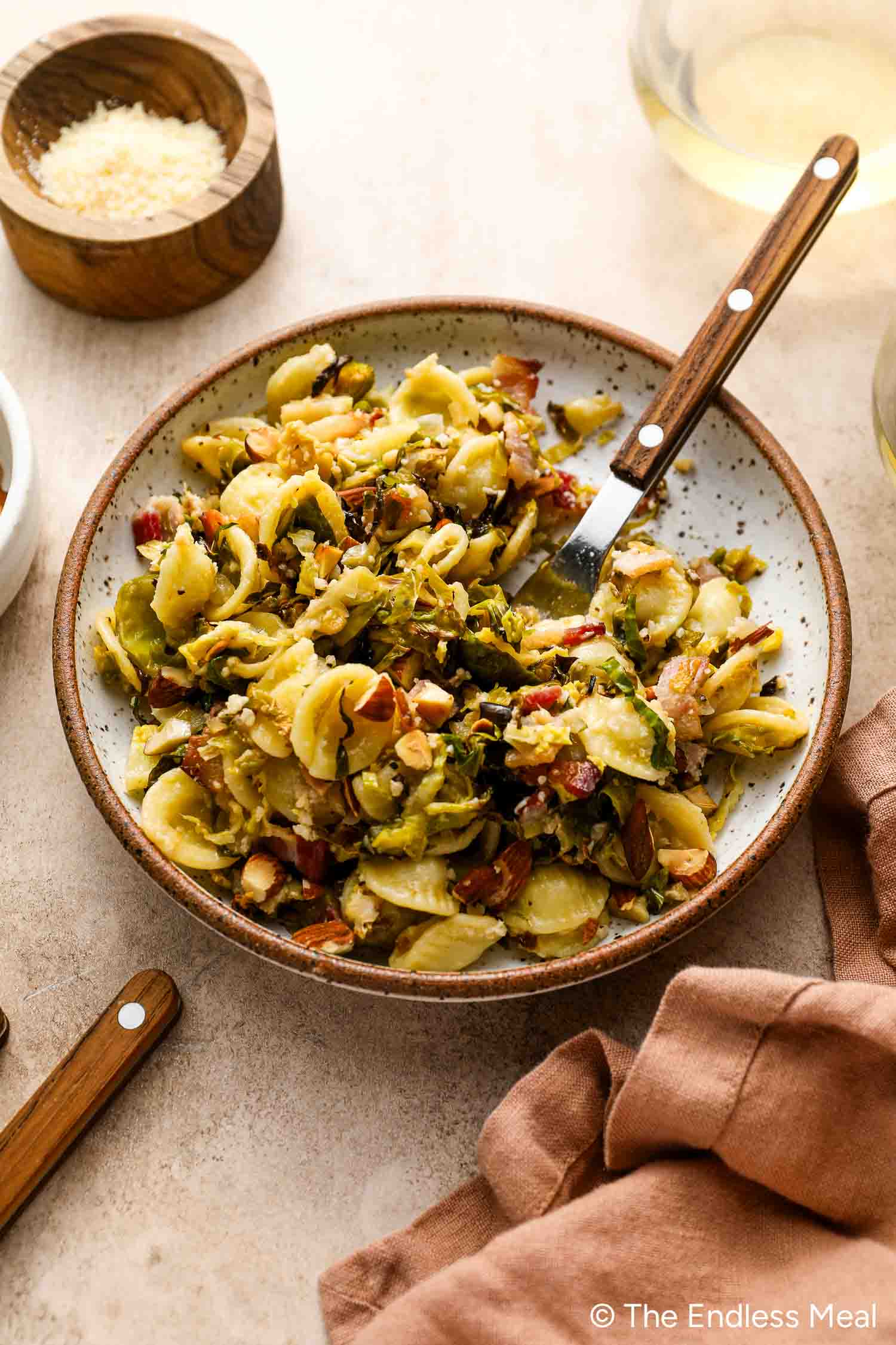A bowl of Roasted Brussels Sprouts Pasta with bacon and chopped nuts sits on the table, accompanied by a fork, wooden bowl of cheese, and a glass of white wine.