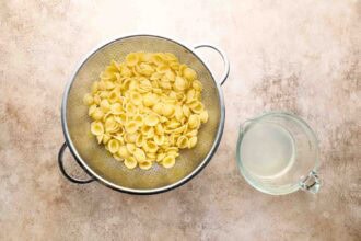 A metal colander filled with cooked orecchiette pasta sits next to a glass measuring cup containing pasta water, ready to be tossed with roasted Brussels sprouts pasta on a beige surface.