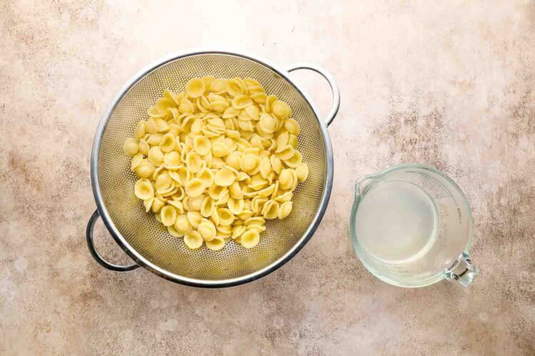A metal colander filled with cooked orecchiette pasta sits next to a glass measuring cup containing pasta water, ready to be tossed with roasted Brussels sprouts pasta on a beige surface.