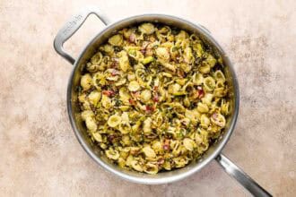 A stainless steel pan filled with Roasted Brussels Sprouts Pasta, featuring cooked orecchiette mixed with vegetables and herbs, sits on a light beige surface.