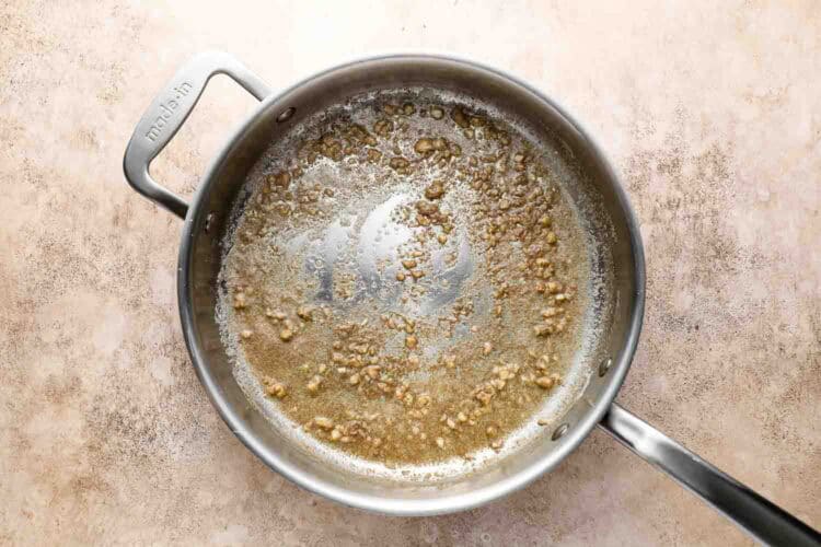 A stainless steel pan with melted butter and minced garlic sautéing, the perfect start for flavorful Pasta or Roasted Brussels Sprouts, on a light textured countertop.