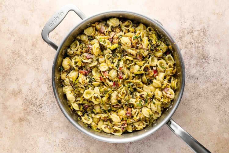 A stainless steel pan filled with Roasted Brussels Sprouts Pasta, featuring cooked orecchiette mixed with vegetables and herbs, sits on a light beige surface.