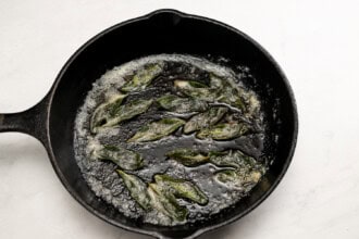 Fried sage leaves sizzling in butter in a black cast iron skillet on a white surface—perfect for topping creamy Pumpkin Tomato Soup.