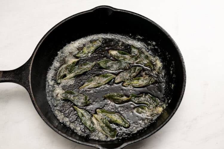 Fried sage leaves sizzling in butter in a black cast iron skillet on a white surface—perfect for topping creamy Pumpkin Tomato Soup.