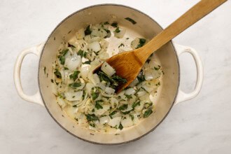 Chopped onions and herbs are being sautéed in a white pot, stirred with a wooden spatula as the base for a flavorful Pumpkin Tomato Soup.