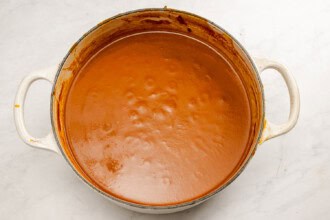 A large white pot filled with smooth, simmering red-orange Pumpkin Tomato Soup sits on a light marble surface.