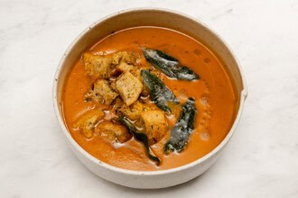A ceramic bowl filled with orange curry, reminiscent of pumpkin tomato soup, featuring pieces of fried bread and garnished with whole green curry leaves, placed on a white surface.