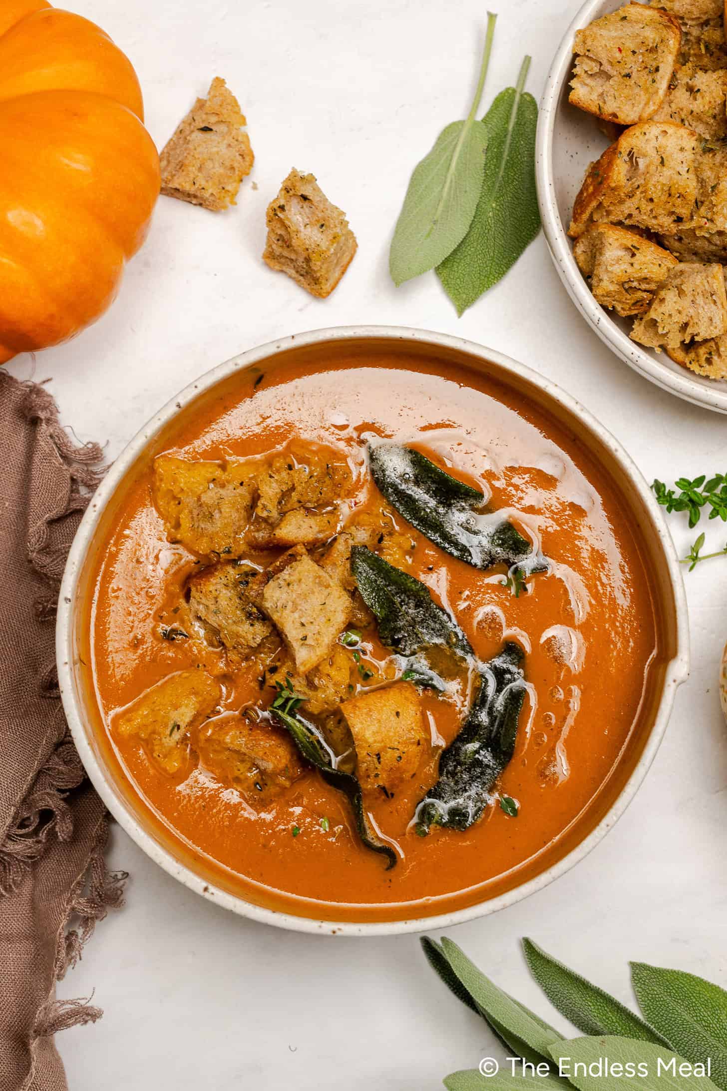 A bowl of creamy Pumpkin Tomato Soup topped with toasted croutons and crispy sage leaves, garnished with fresh herbs and a small pumpkin nearby.
