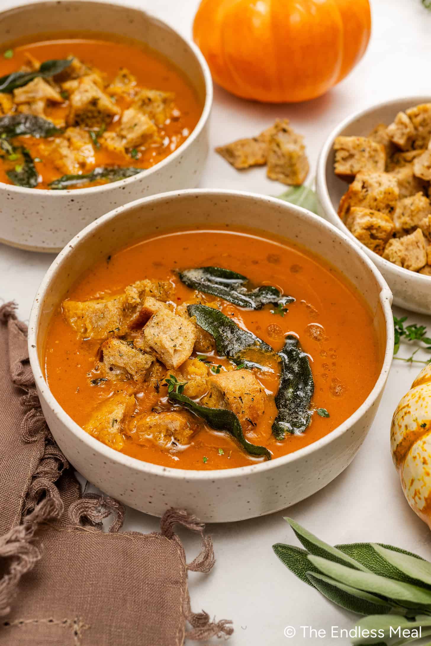 A bowl of creamy pumpkin soup topped with croutons and crispy sage leaves, with another bowl, a pumpkin, and a small dish of croutons nearby.