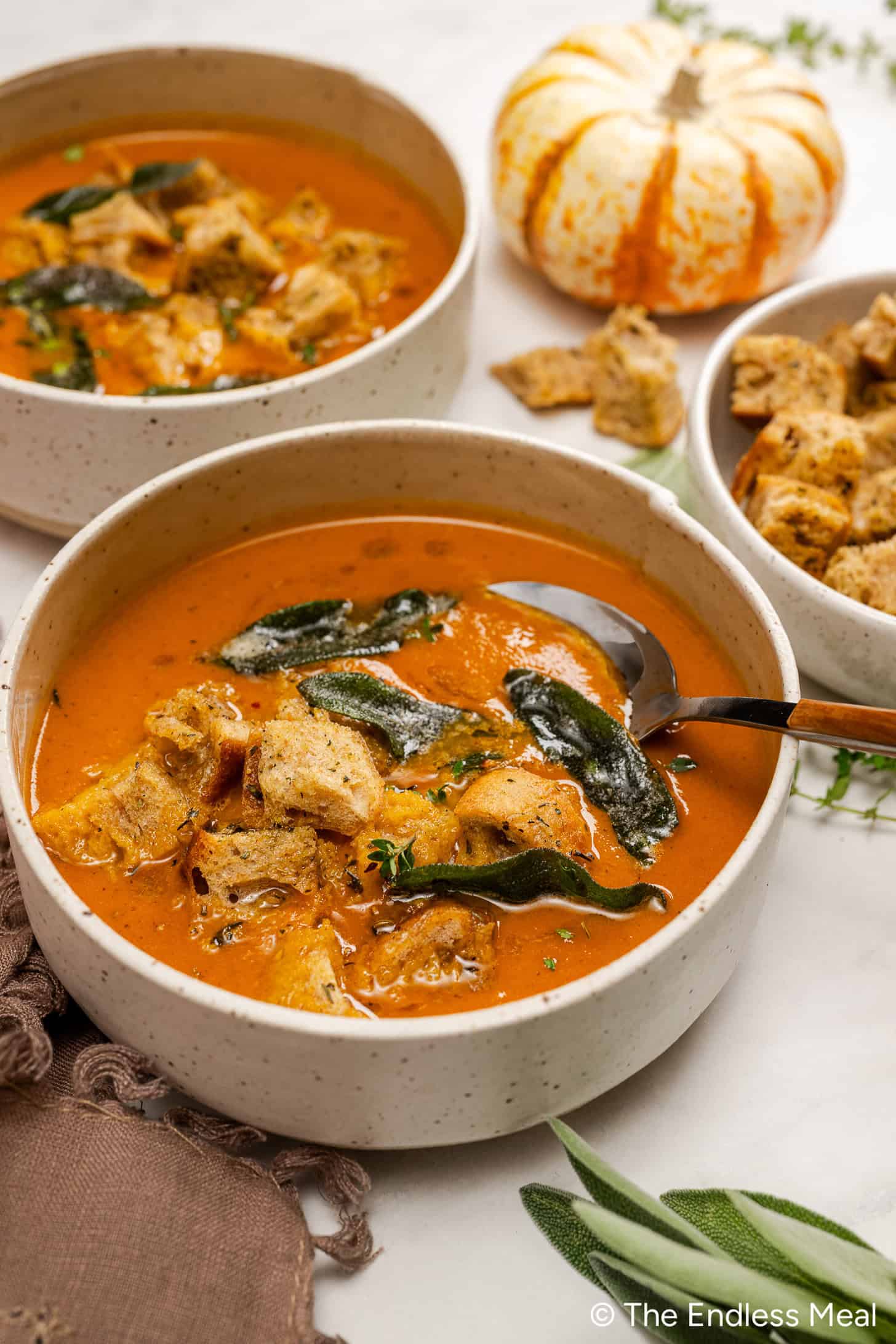Two bowls of creamy Pumpkin Tomato Soup topped with croutons and crispy herbs sit on a white surface, accompanied by a small decorative pumpkin and a bowl of extra croutons.