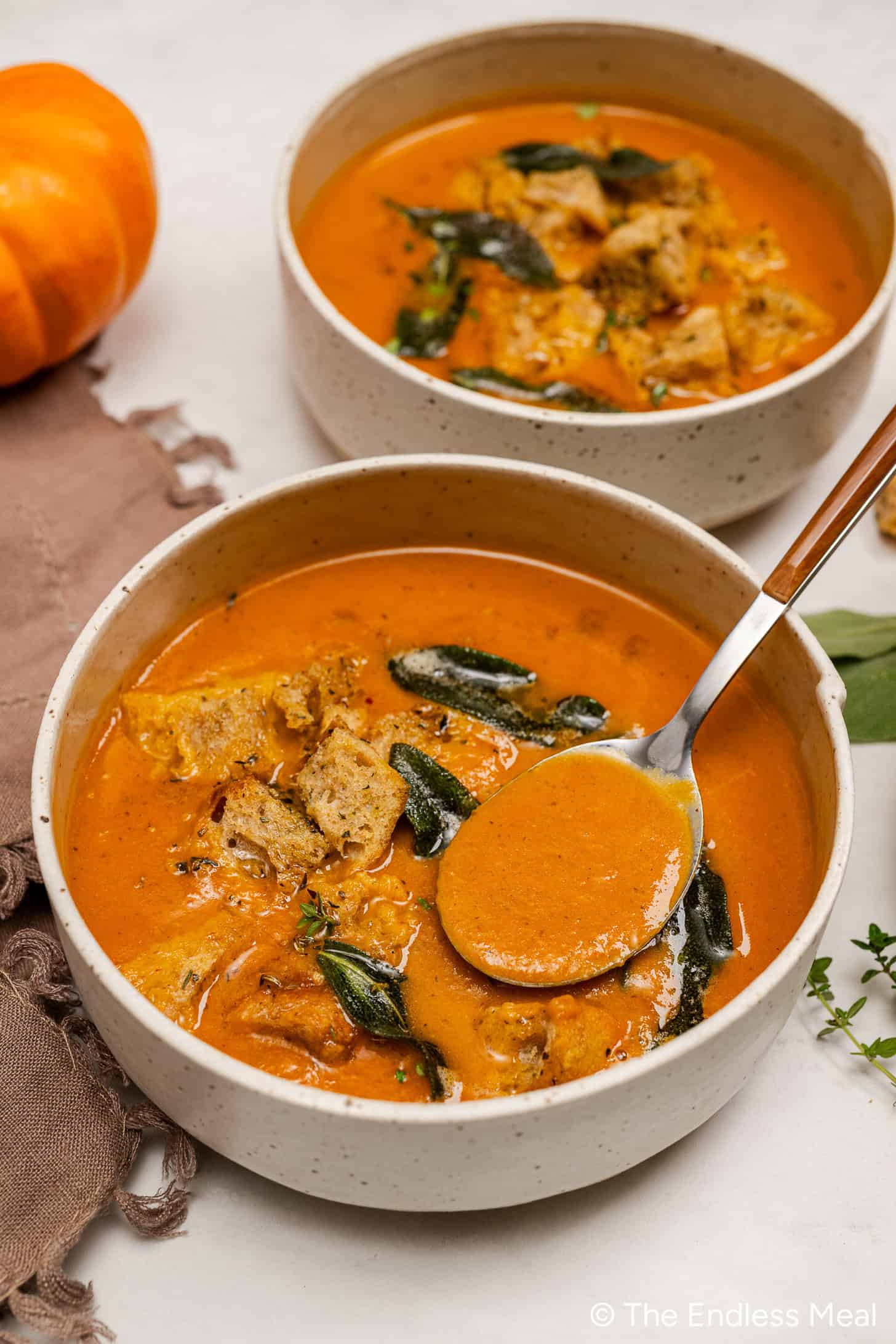 Two bowls of creamy Pumpkin Tomato Soup, garnished with croutons and herbs, sit on a white surface. A spoon hovers above one bowl, while a small pumpkin and napkin add a cozy touch nearby.