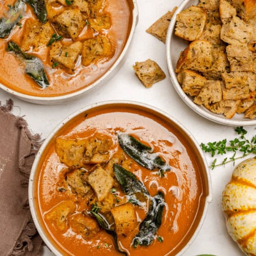 Two bowls of creamy Pumpkin Tomato Soup topped with croutons and herbs, with an extra bowl of croutons, pumpkins, and fresh sage on a white surface.