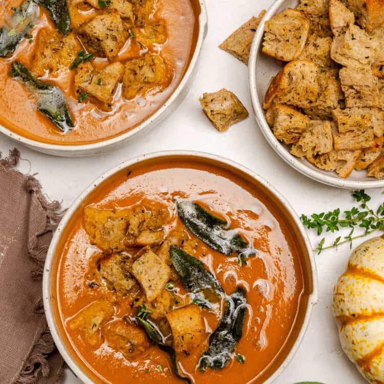 Two bowls of creamy Pumpkin Tomato Soup topped with croutons and herbs, with an extra bowl of croutons, pumpkins, and fresh sage on a white surface.