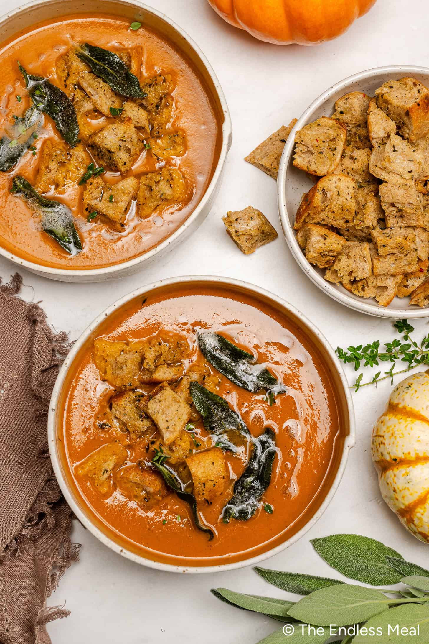 Two bowls of creamy Pumpkin Tomato Soup topped with croutons and herbs, with an extra bowl of croutons, pumpkins, and fresh sage on a white surface.