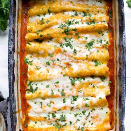 A casserole dish filled with baked chicken enchiladas topped with melted cheese and chopped herbs, sitting on a white surface.