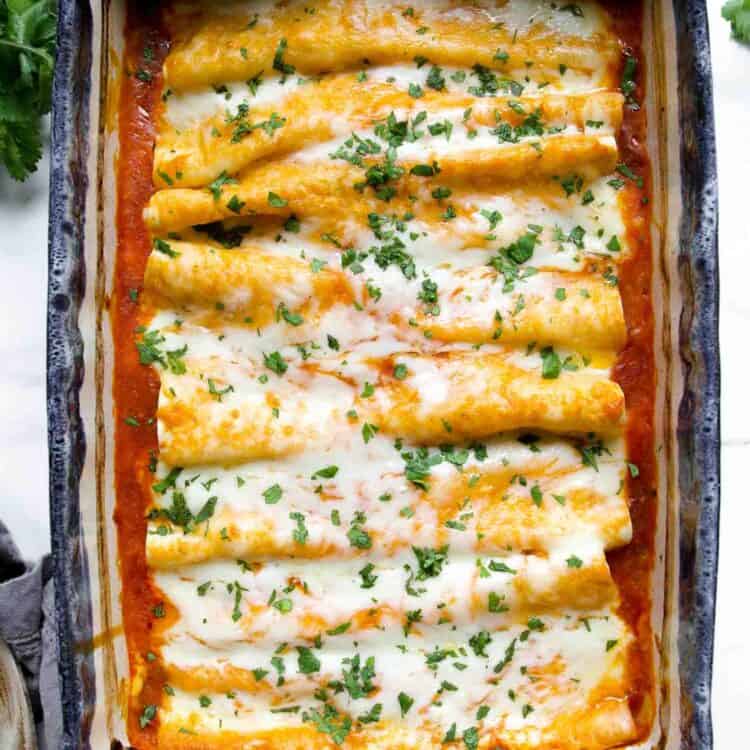 A casserole dish filled with baked chicken enchiladas topped with melted cheese and chopped herbs, sitting on a white surface.