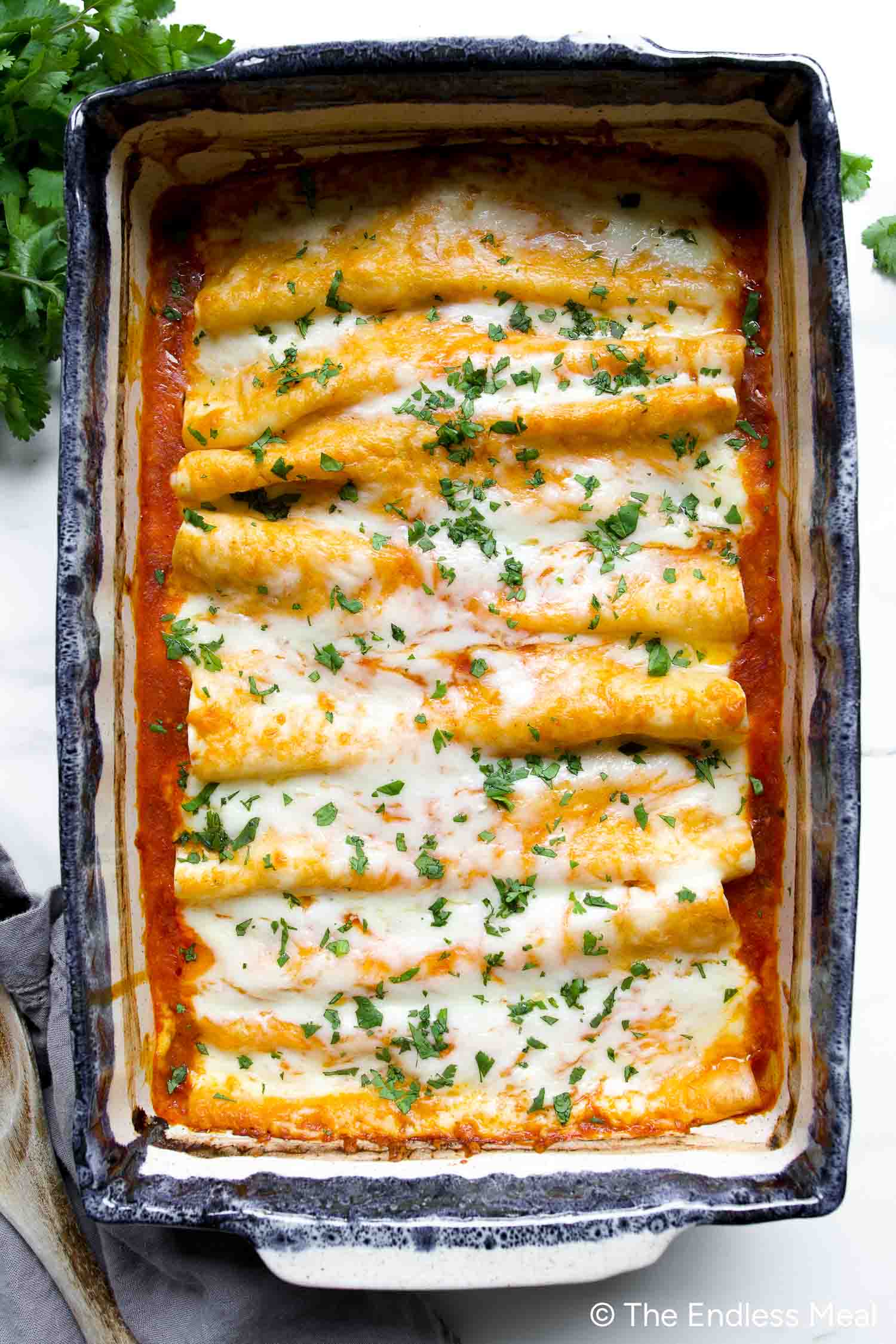 A casserole dish filled with baked chicken enchiladas topped with melted cheese and chopped herbs, sitting on a white surface.