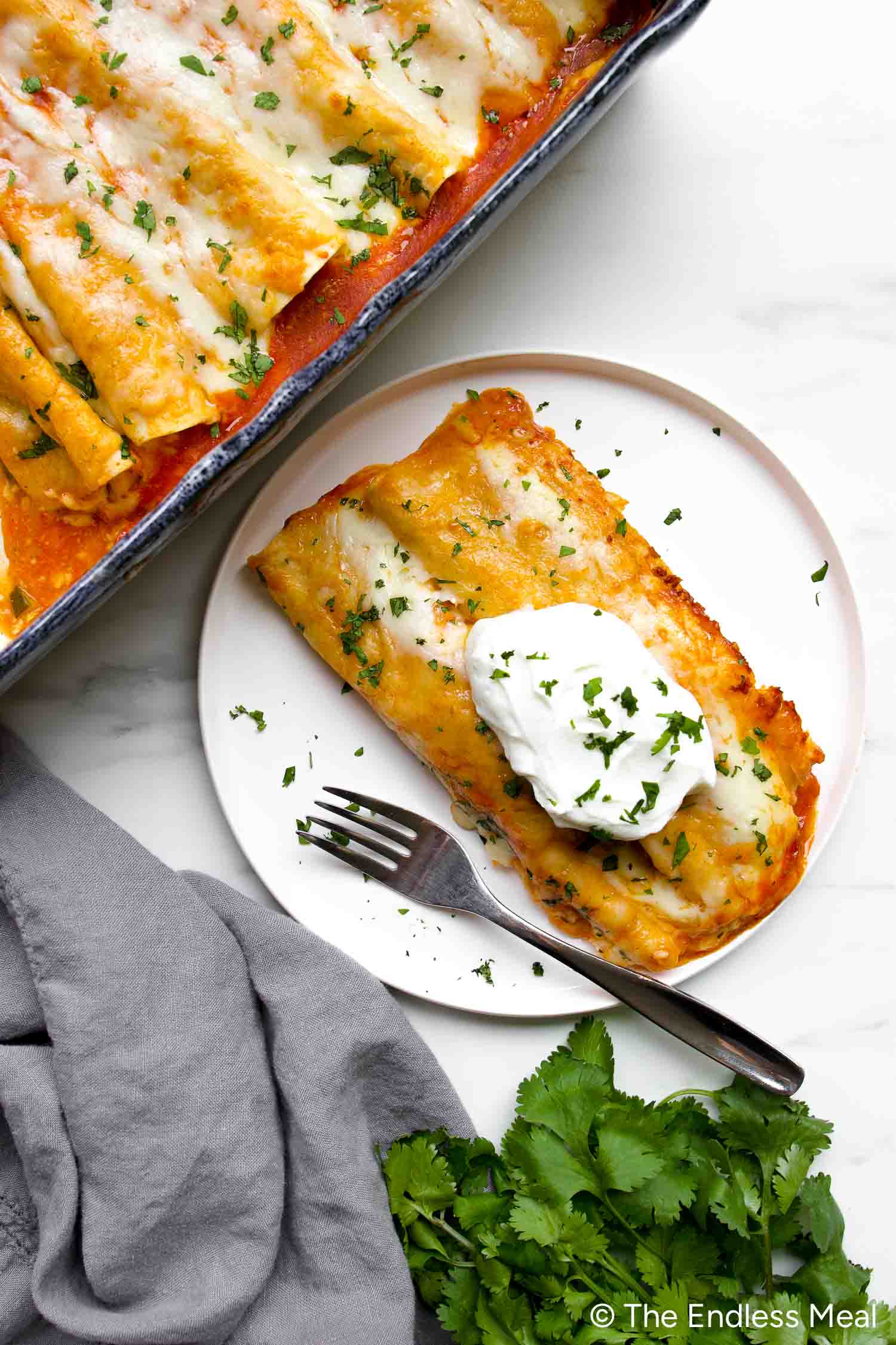 A plate with two Chicken Veggie Enchiladas topped with sour cream and chopped cilantro sits next to a baking dish of more enchiladas. A fork and fresh cilantro are beside the plate.