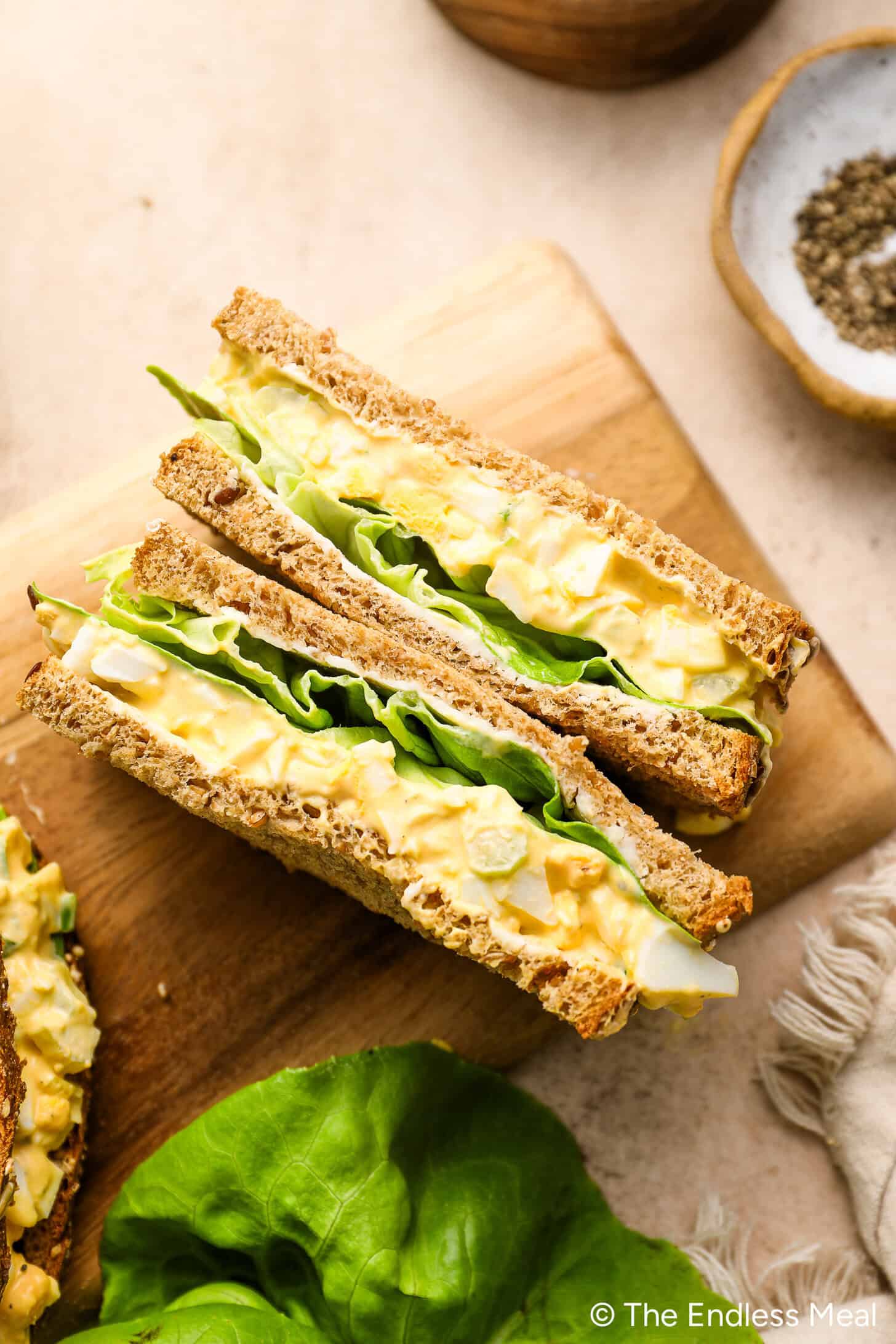 Egg salad sandwich with lettuce on whole grain bread, sliced in half and placed on a wooden cutting board.