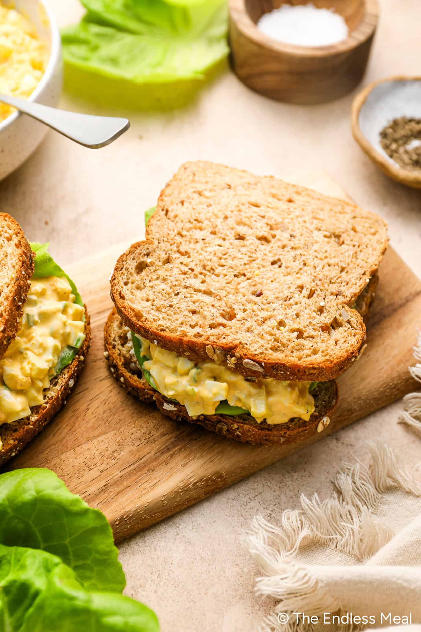 Egg salad sandwich with lettuce on whole grain bread, placed on a wooden board, with a bowl of seasoning and lettuce leaves in the background.
