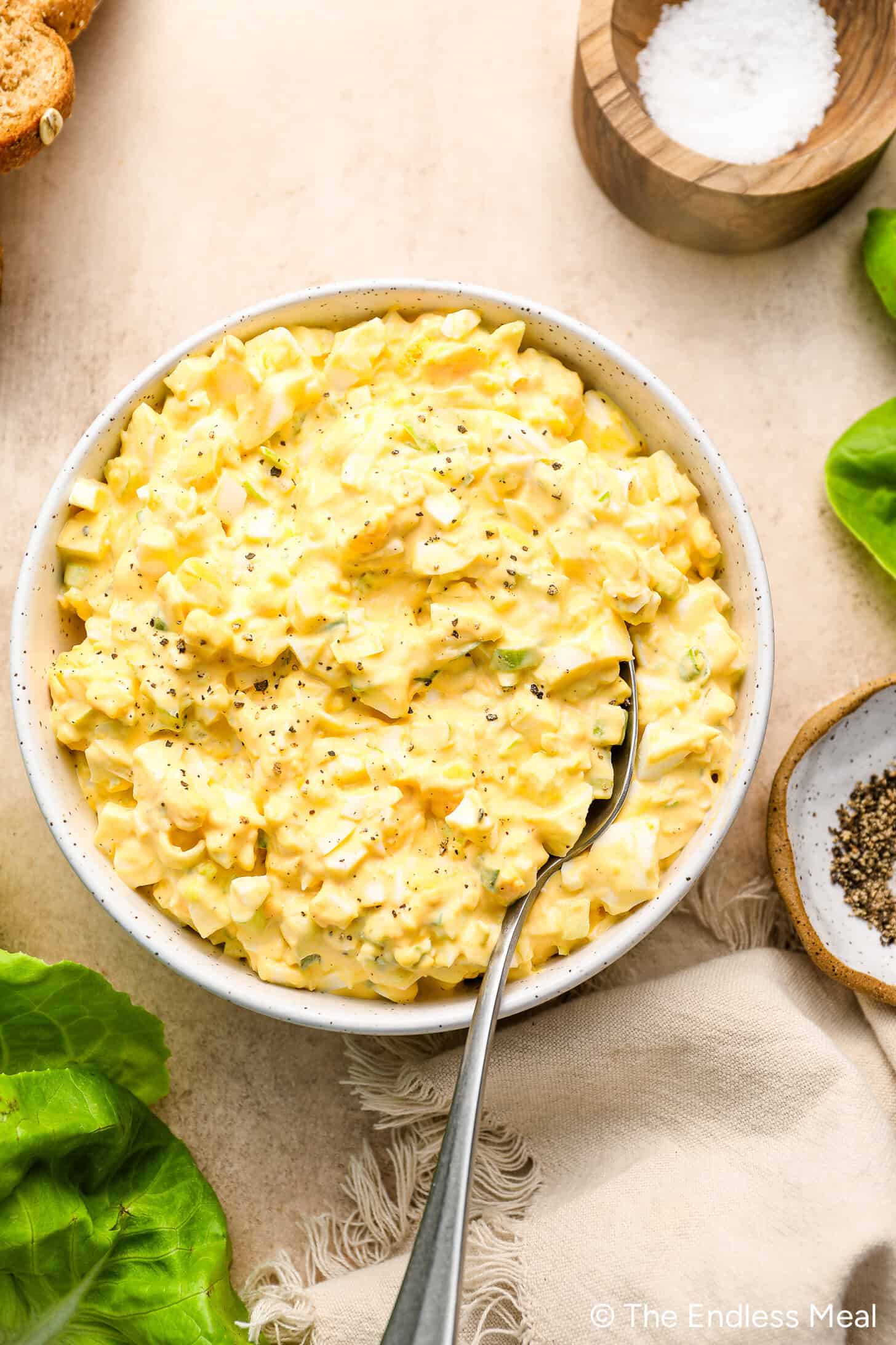 A bowl of egg salad with chopped eggs and celery, garnished with black pepper, sits on a beige surface next to salt, pepper, lettuce leaves, and a spoon.