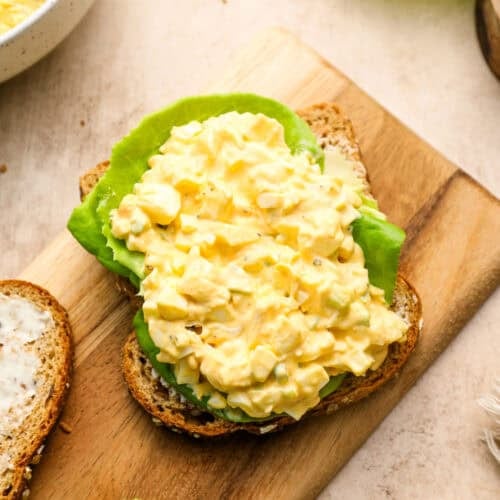 Open-faced egg salad sandwich with lettuce on whole grain bread, placed on a wooden board. A bowl of egg salad and lettuce leaves are nearby.
