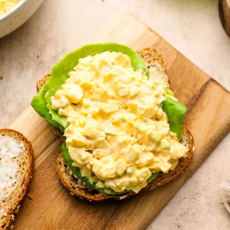 Open-faced egg salad sandwich with lettuce on whole grain bread, placed on a wooden board. A bowl of egg salad and lettuce leaves are nearby.